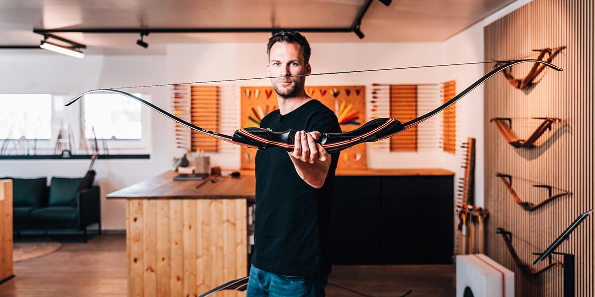 BEARPAW Store Rossach – Experte präsentiert einen hochwertigen Langbogen, umgeben von Bogenkollektion an der Wand, ideal für Beratung und Tests nahe Coburg.